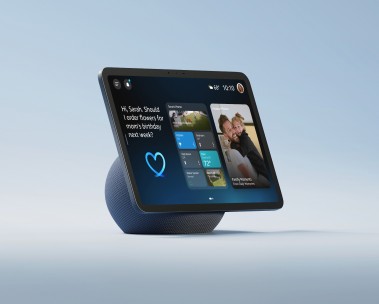 Smart display with blue base showing digital assistant message: "Hi, Sarah. Should I order flowers for mom's birthday next week?" with heart icon, weather widget and family photos.