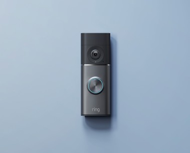A Ring doorbell mounted on a wall. The device has a dark gray/black design with a camera lens at top and circular button with blue illuminated ring at bottom. 