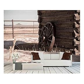 A Wagon Wheel Resting Against an Old Cabin with Mountains in The Distance - Removable Wall Mural | Self-Adhesive Large Wallpaper - 66x96 inches