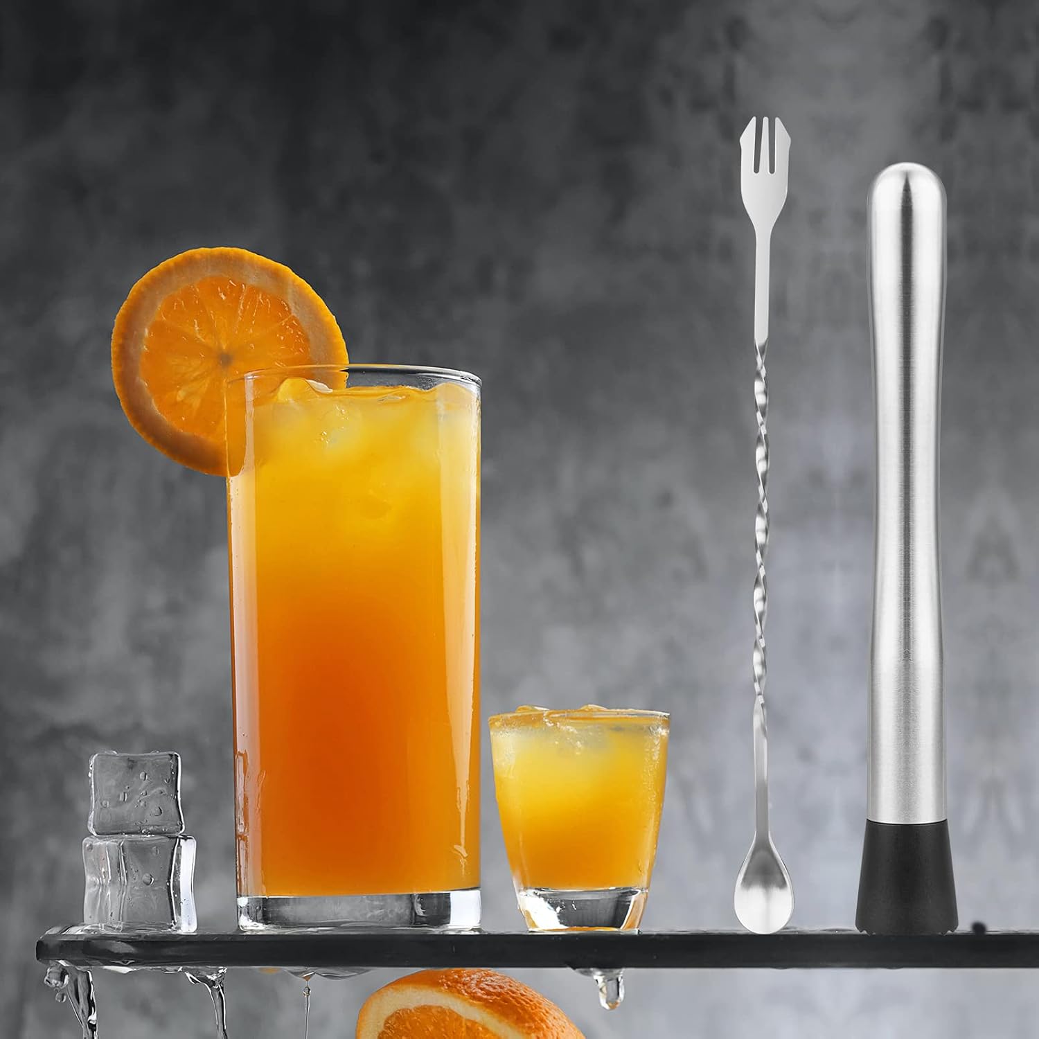 stainless steel muddler and spiral bar spoon standing upright beside two tall orange drinks on a grey concrete surface