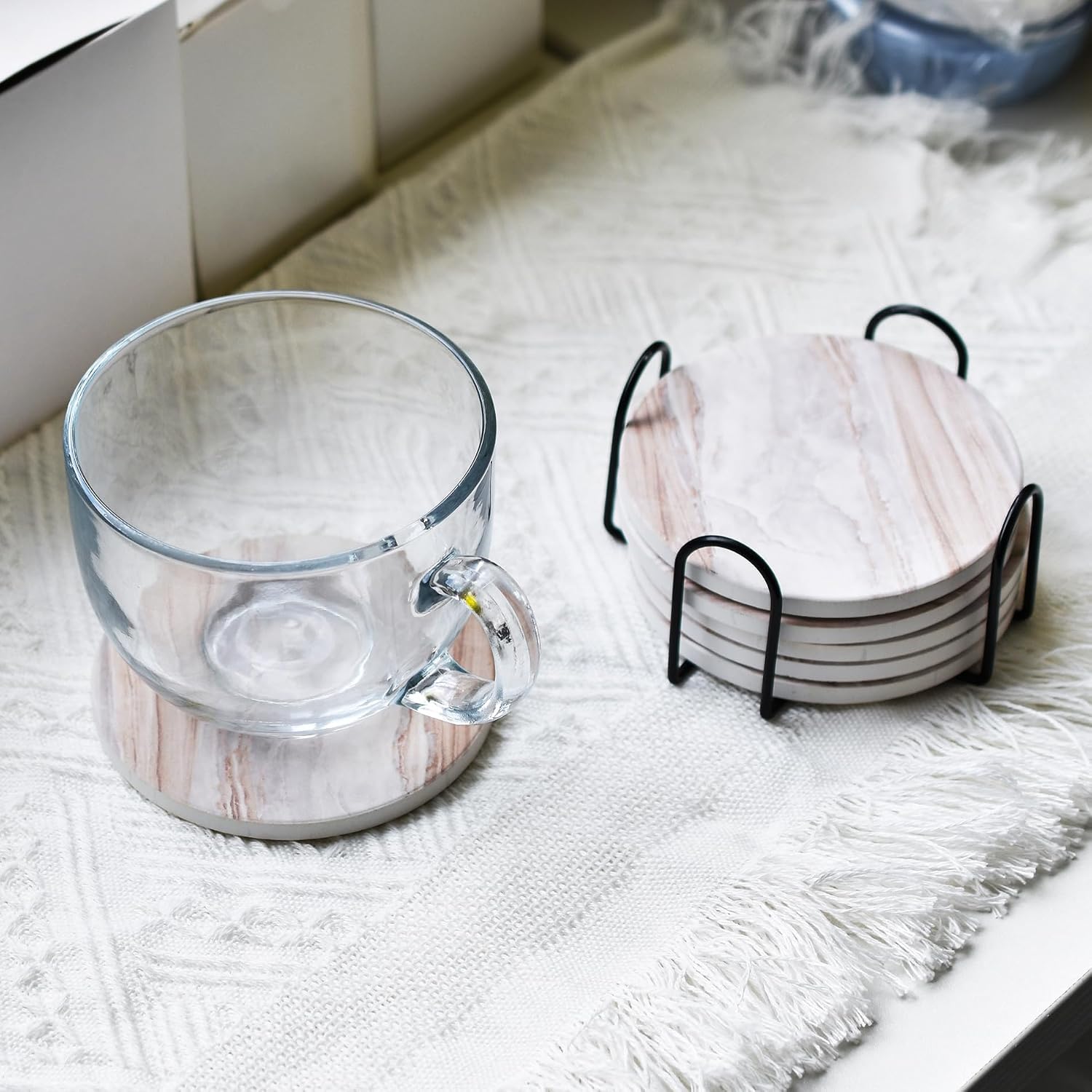 marble-patterned ceramic coasters stacked in a black metal holder on a marble counter with a glass pitcher behind