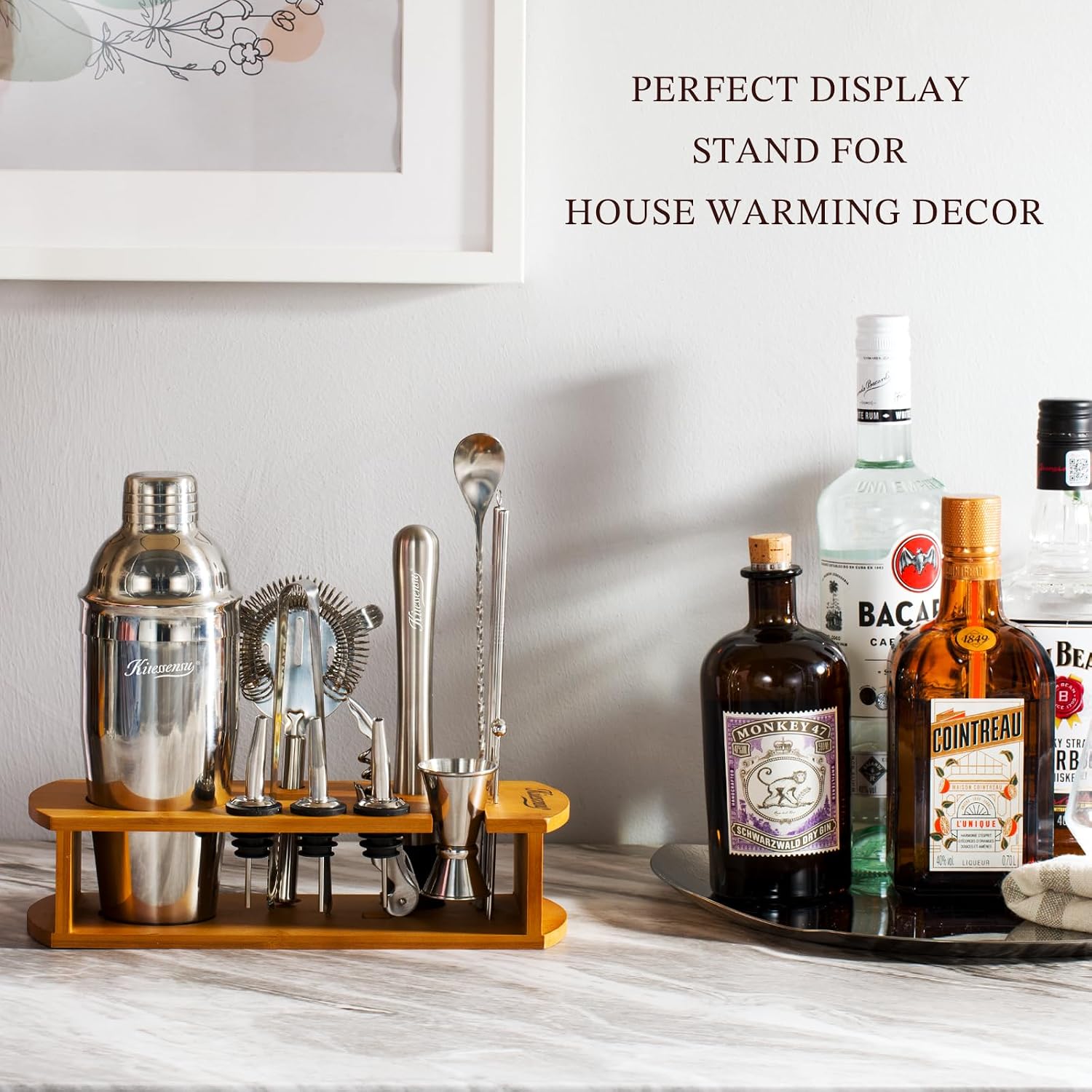 stainless bartender kit arranged on a bamboo stand on a marble counter with spirit bottles behind it and framed art on the wall