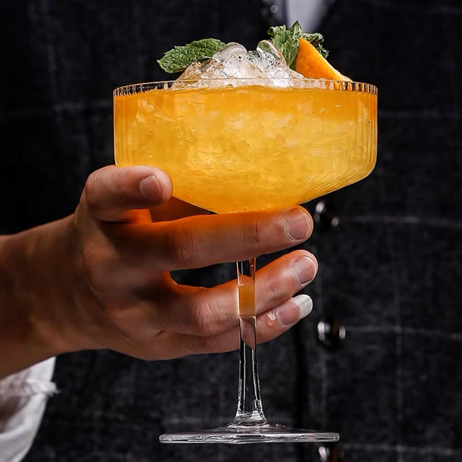 hand holding a ribbed coupe glass with a garnished amber cocktail in it against a warm dark out-of-focus room