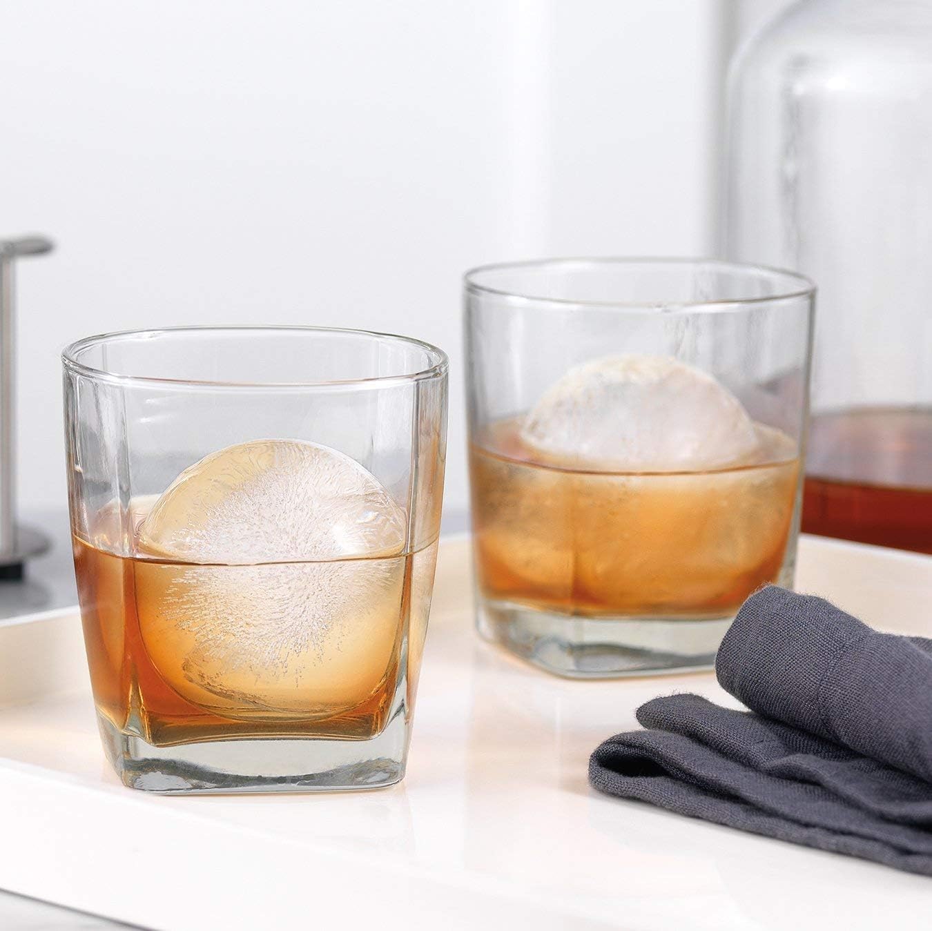 round silicone ice sphere mold on a pale kitchen counter beside two rocks glasses each holding a large ice sphere and an amber pour