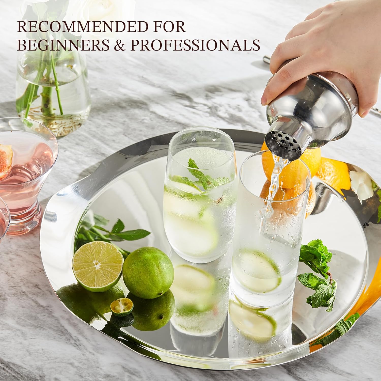 hands pouring from a stainless cocktail shaker into a tall iced drink on a silver tray set with limes and mint