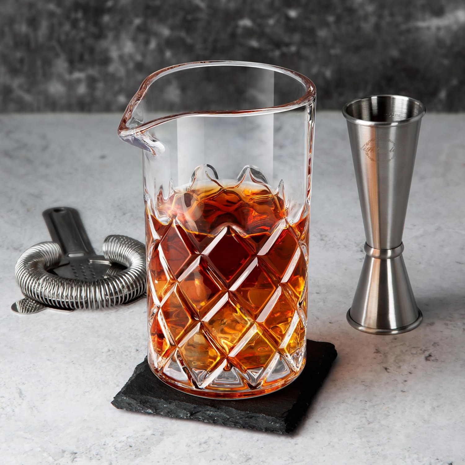 cut crystal mixing glass holding a stirred amber drink on a slate coaster against a pale stone wall with a jigger and strainer alongside