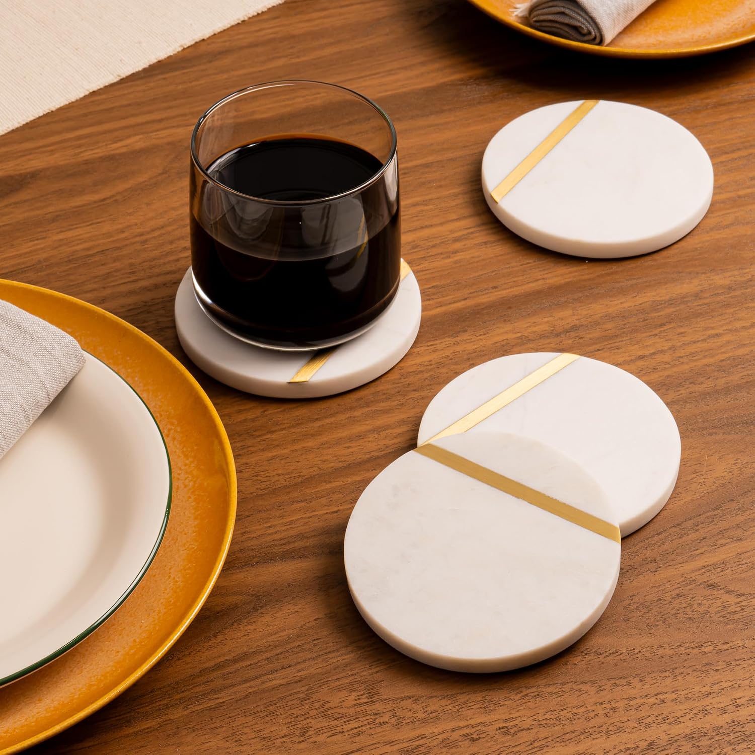 two white marble coasters with gold inlay on a warm wood table, one holding a filled rocks glass beside gold-rimmed plates