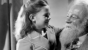 Edmund Gwenn as Kris Kringle in his Santa Claus outfit looking at a little girl, in the Miracle on 34th Street game